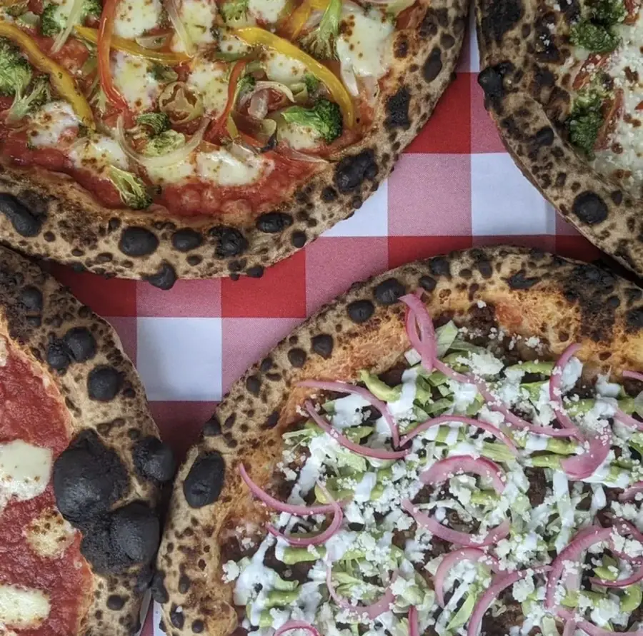 Gourmet Neapolitan wood-fired pizzas on a red and white checkered tablecloth. Features a vibrant vegetable pizza with bell peppers and broccoli, and a unique Mexican-style pizza topped with chorizo, pickled red onions, avocado, lettuce, and crumbled cheese. Ideal for brewery and restaurant menus.