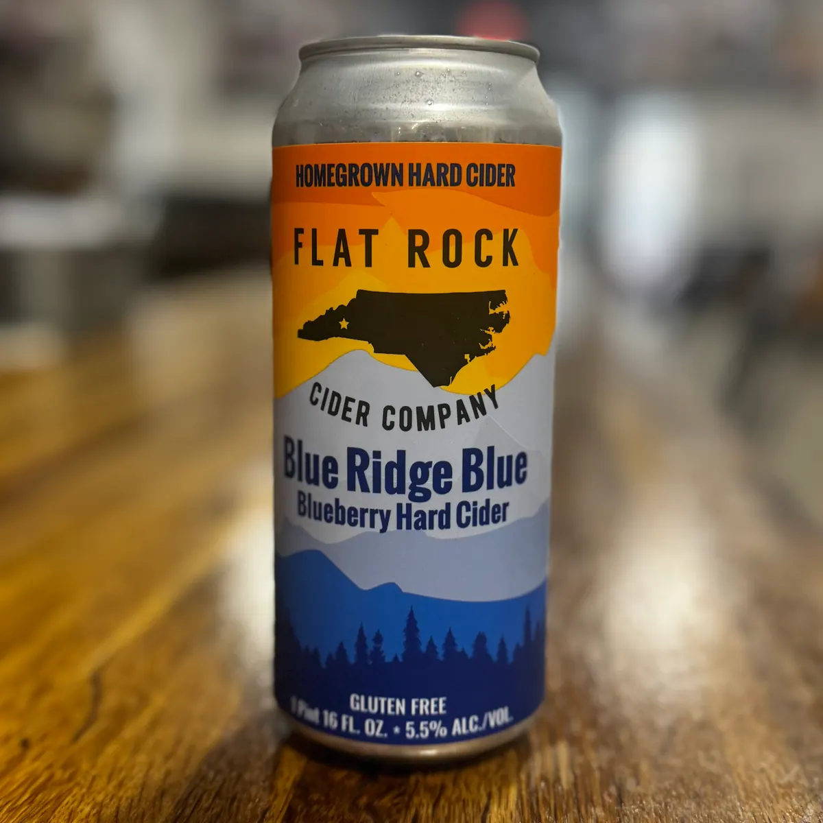 A 16 oz can of Flat Rock Cider Company Blue Ridge Blue Blueberry Hard Cider, a gluten-free homegrown hard cider from North Carolina, sitting on a wooden bar top.